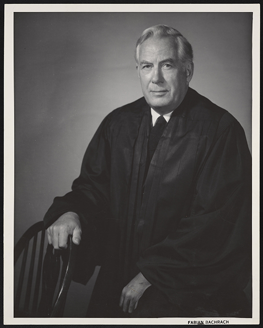 A man in a black robe sits next to a chair, with his right hand resting on it.
