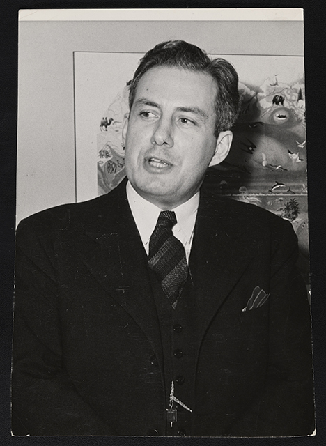 A standing man wearing a suit and tie, shown from the waist up to his head. Behind him is a piece of art hanging on a wall. 
