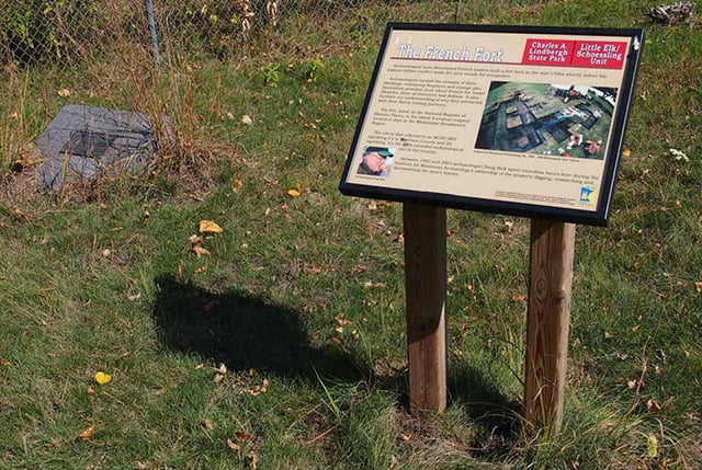A sign with text and a color image stands on two wooden supports in front of a grassy background. A historical marker set into the earth is visible at left.