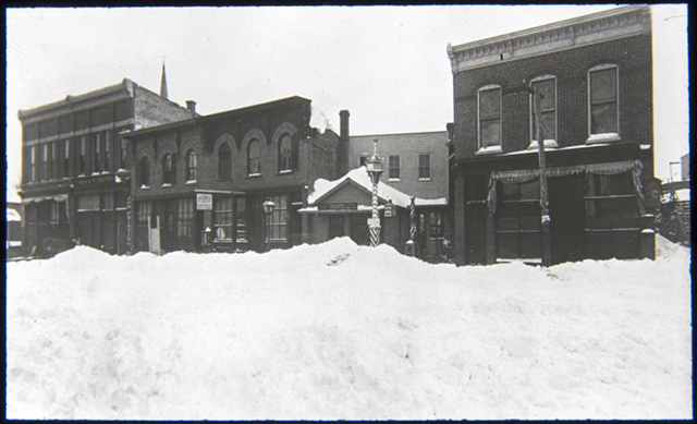 Four buildings line a wintery street scene, with snow piled up in the foreground.