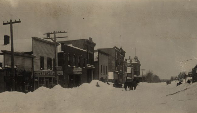 Piles of snow line a street with buildings lined up on the left and right sides, with telephone poles marking both.
