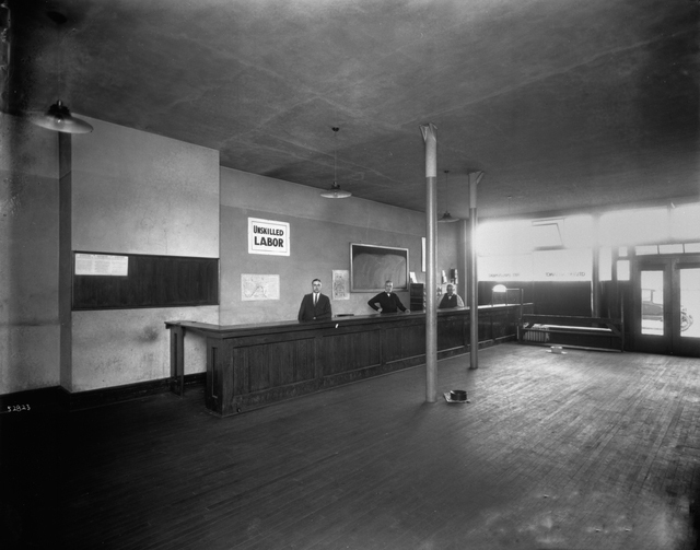 A mostly empty room with a wooden floor. Four men stand behind a countertop at the center.