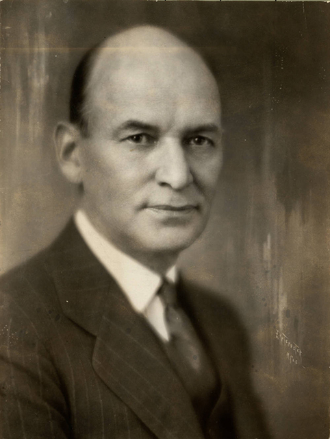 The head and shoulders of bald man wearing a collar, tie, and suit jacket.