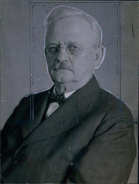 Head and shoulders of a man with a moustache, glasses, and bow tie.