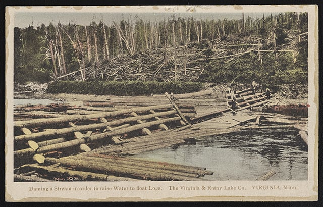 A dammed stream with logs and dam components covering most of the water. Leafless trees stand on the bank in the background.