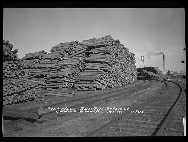 Two railroad tracks curve to the left of the image around stacks of logs, with a smokestack and water tower in the background.