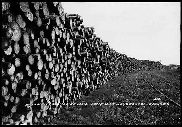 Stacks of wood fill the left-hand side of the image and extend across the picture plane to a vanishing point at right. Railroad tracks run through the grass at center right.
