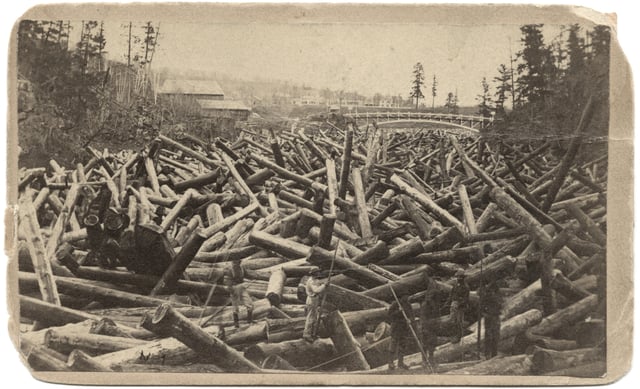 Laborers stand on jammed logs in the foreground, with logs continuing to a bridge in the background. Trees stand in the right and left background.