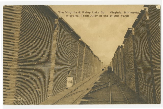 Railroad tracks run through an alley created by two rows of stacks wooden boards at the left and right, rising up to a sepia-colored sky.