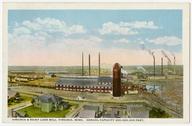Bird's-eye view of a lumber mill showing a red multistory building with a red silo next to it, smaller buildings in the foreground, and smokestacks in the background, all under a blue sky with white clouds.