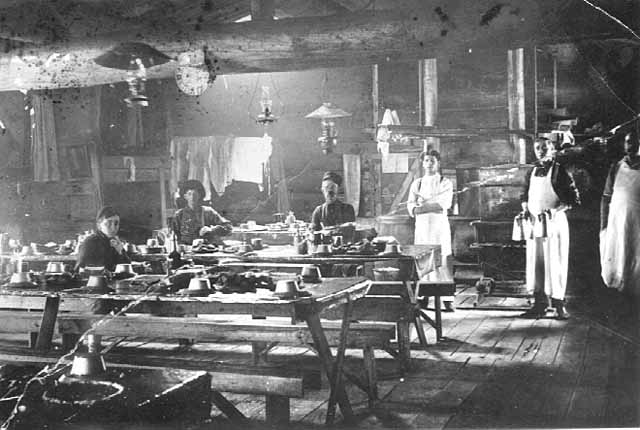 Three people sit and three others, wearing aprons, stand inside a dining hall with tables, benches, and an exposed wooden rafter overhead.