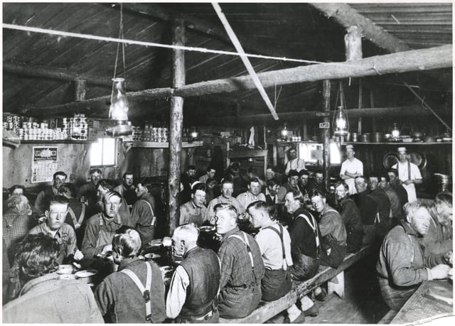 The interior of a lumber camp building showing three long tables with laborers sitting on benches. The building's rafters are visible above them.