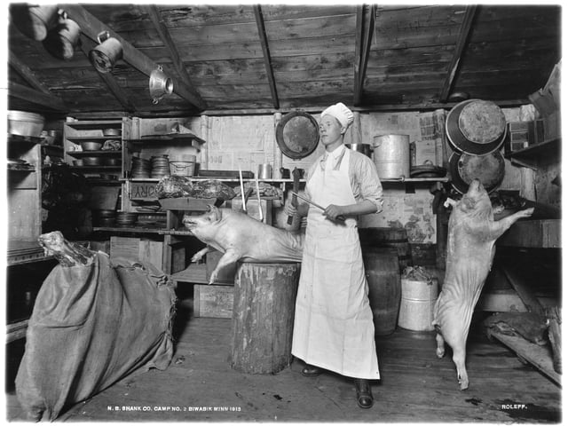 A man in a white apron and cook's hat stands between two slaughtered pigs with knives in his hands. He stands inside a kitchen with a wooden roof.
