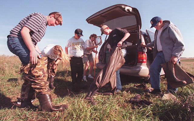 Six people stand grouped together in front of a minivan with its trunk door open. Three of the people prepared to put on waders..