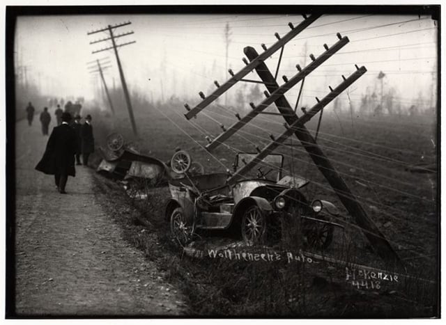People in white collars, hats, and long coats walk along a road at right, with burned cars and leaning power lines at right.