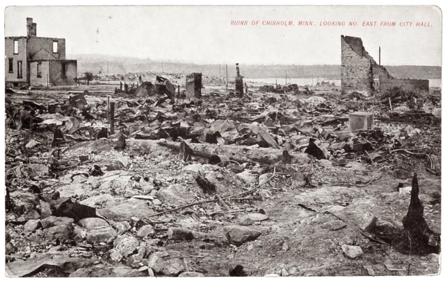 Rocks, charred tree stumps, and building ruins stretch to a distance horizon with a lake to the right. The walls of two burned buildings still stand.