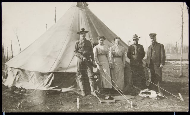 Three men (two of them in military uniform) and two women post in front of a round canvas tent, with the charred remains of trees to the right and left.