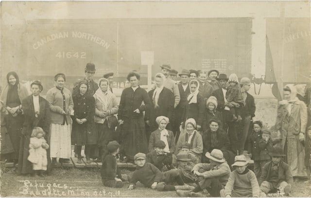 A crowd of people stands in front of a Canadian Northern train car. Women and children cluster in the front, with men closer to the back. They wear hats, kerchiefs, shawls, and coats.