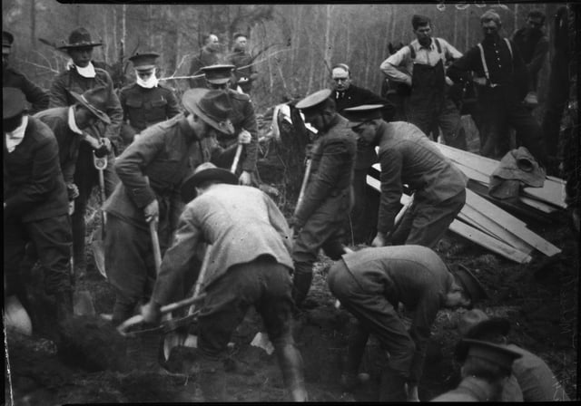 Men in military uniforms holding shovels stand around a circle of earth, with planks of wood to their right and more people and forest behind them.