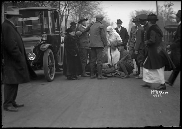 A man watches from the left foreground (an automobile stands behind him) watching a group of aid workers attending to a fire victim or victims.