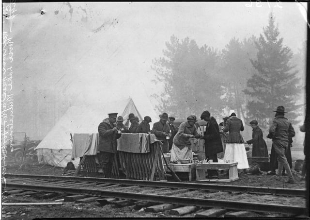Behind a train track and in front of a stand of trees, workers distribute food and drinks to men in uniforms.