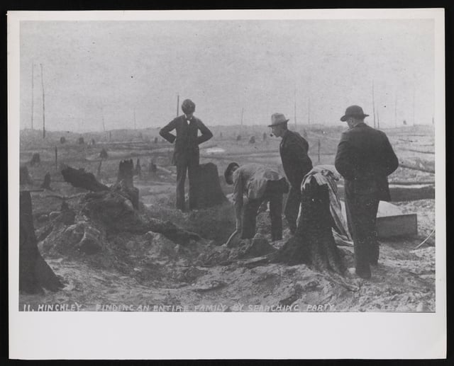 Four men wearing hats gathering around a hole in charred earth, with burnt tree stumps close to them as well as in the background.