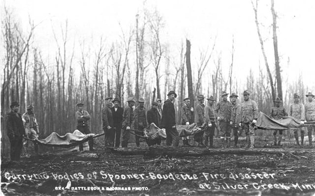 About twenty men, some wearing military uniforms, pose in front of burned, leafless trees. Some of them carry stretchers that lift up the bodies of three fire victims.