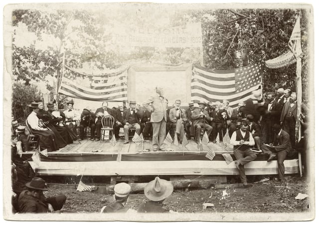 Stage platform with observers in front and people sitting in chairs in front of them, with a speaker standing front and center.