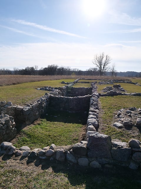 Stone foundation ruins in a grassy field, with trees and sky in the background.
