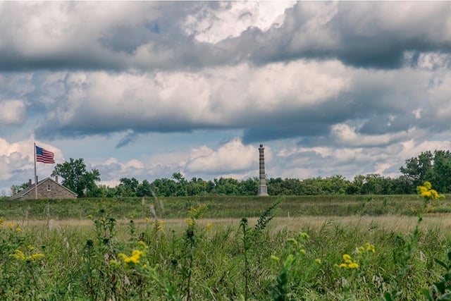 Grassy foreground with yellow flowers below a blue but cloud-filled sky, with a flag at left and a pillar at center.