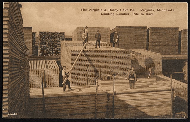 Stacks of lumber rise to the sky in stacks on two different levels above the ground, with workers posed on on each of them.