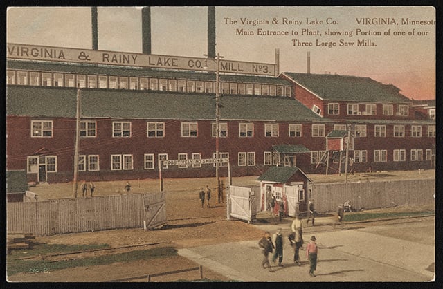 Four-story lumber mill building with a red facade, green roofs, and white fence in the middle ground.