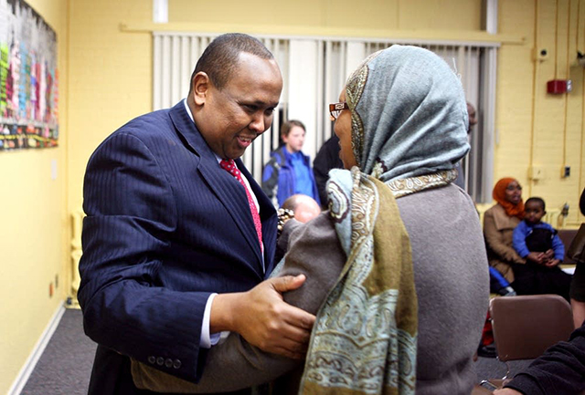 A man in a blue suit jacket holds the left arm of a woman wearing hijab, with yellow walls and other people behind them.