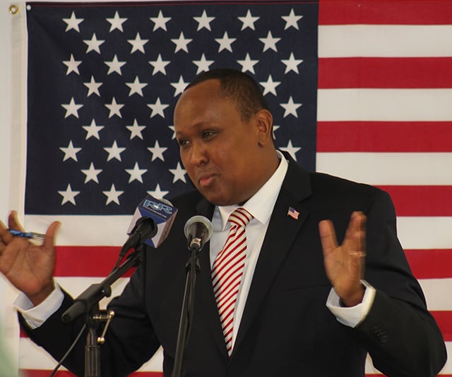 A man wearing a red-and-white-striped tie, a blue suit jacket, and a white dress shirt stands in front of a microphone with his hands lifted. An American flag is behind him.