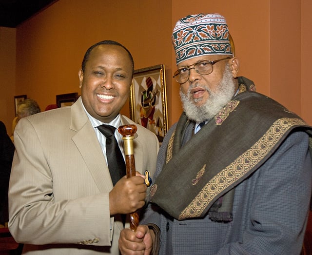 Two men stand next to each other. One wears a tan suit and a black tie and smiles broadly. The other wears a blue suit jacket, a textile wrap around his shoulders, and a kofia on his head. Both men grasp a wooden stick betwen them.