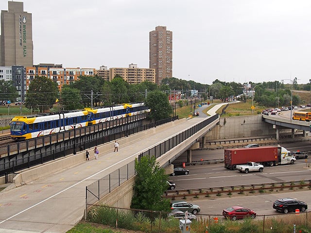 A light-rail train crosses a bridge over a freeway as vehicles pass underneath.