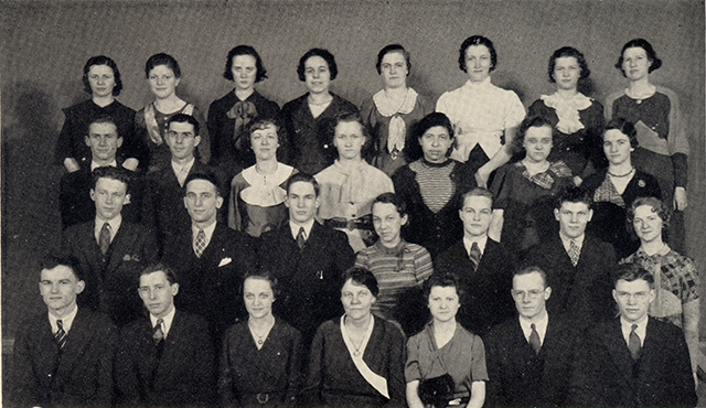 Students stand in four staggered rows of six or seven people each. They wear formal dresses and suits.