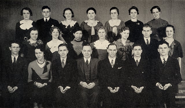 Students stand in three staggered rows of seven people each. They wear formal dresses and suits.