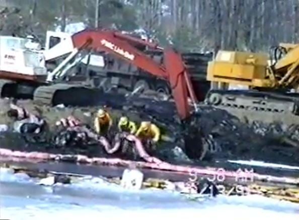 Workers in yellow protective gear handle a pink pipeline in front of construction vehicles, with a frozen stretch of water in the foreground.