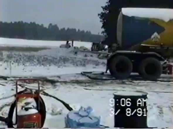 Part of an oil tanker is visible on the right side of the image, with hoses snaking around it. Snow covers the foreground to the background, with a tree line behind that. 