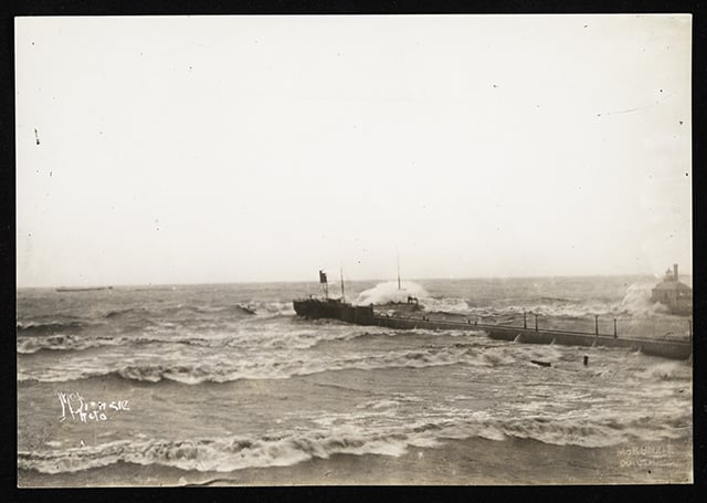 Waves crash toward a beach on two sides of a pier, with a block of white sky above.