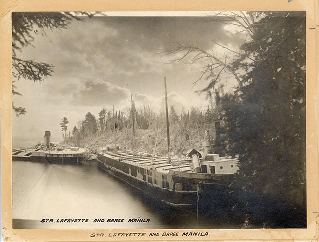 A damaged ship sits in water close to a shoreline wrecked by wind, with a second boat behind it.