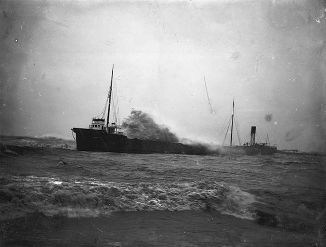 A wave meets a sandy shore, while behind it a ship moves through choppy waves.