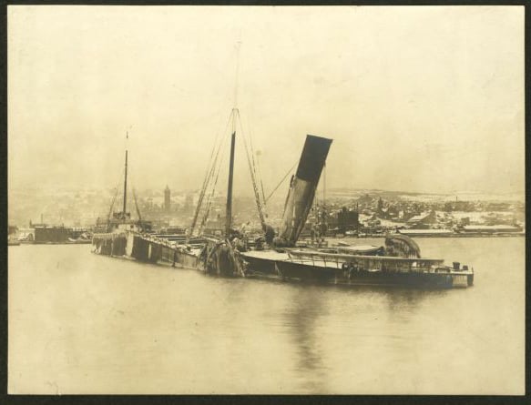 A wrecked steamship sits partially submerged in water, with its bow facing the shore of a lake, with buildings behind it.