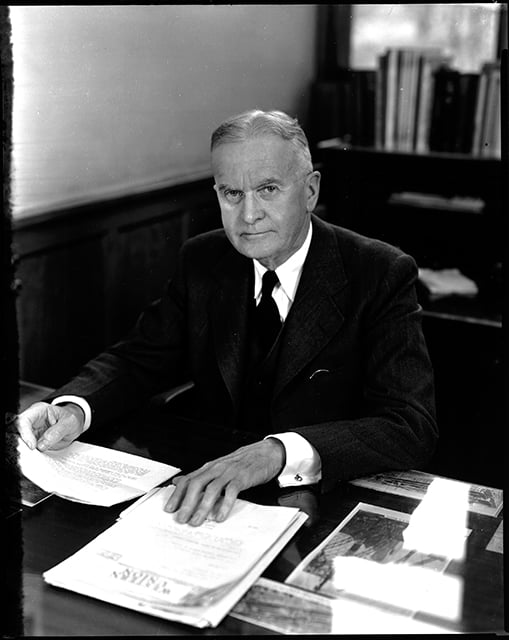 Man in a white dress shirt, tie, and suit jacket sitting at a desk with papers.
