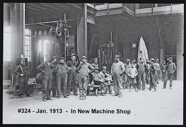 Adults and children stand in an open space in front of a factory building.