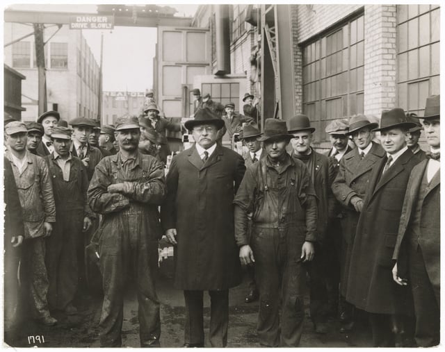 Men wearing caps and brimmed hats pose in front of factory buildings.