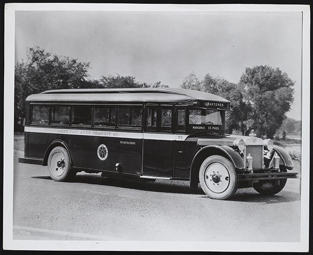 1920s-era bus viewed at a shallow three-quarter angle in front of a tree line with sky in the background