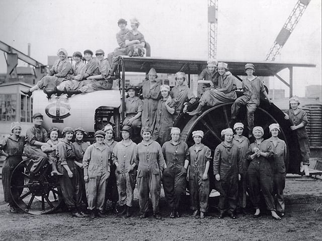 Women workers waring hats and overalls pose in front of a tractor.