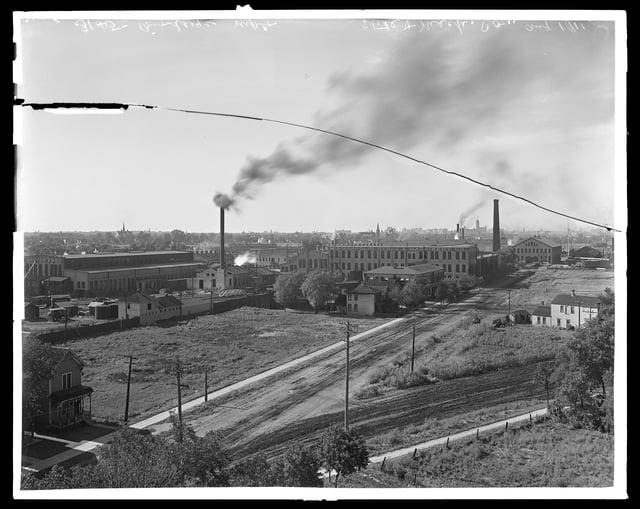 Bird's eye view of a factory, in the middle ground, with smoke billowing from a stack and a grey sky above them in the background.